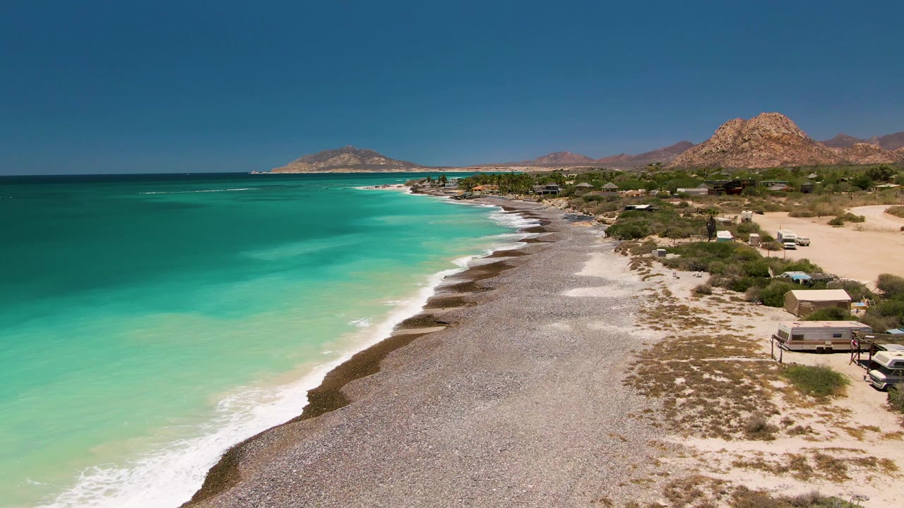 Cabo Pulmo shoreline from above with a wider band of pale surf against the pebbles, turquoise ocean to the left, small structures to the right, and no visible people.