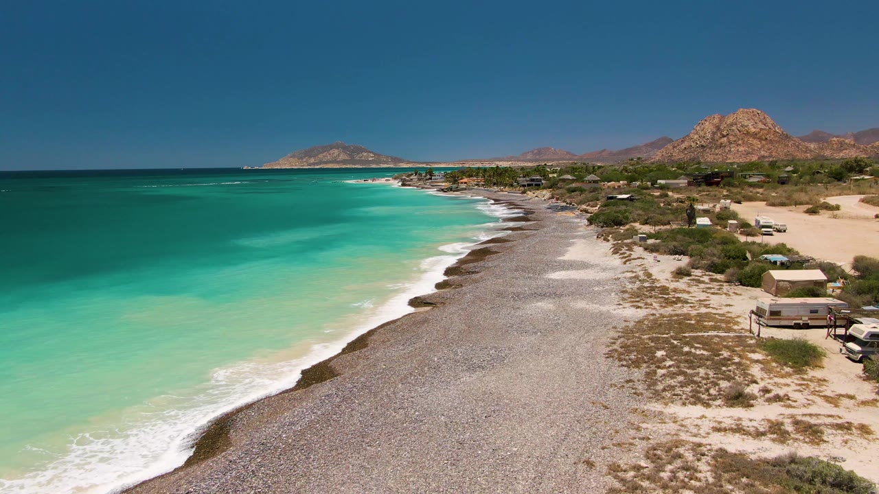 Aerial frame of the Cabo Pulmo coast with the beach curving toward distant hills, green-blue water on the left, shore structures on the right, and no visible people.