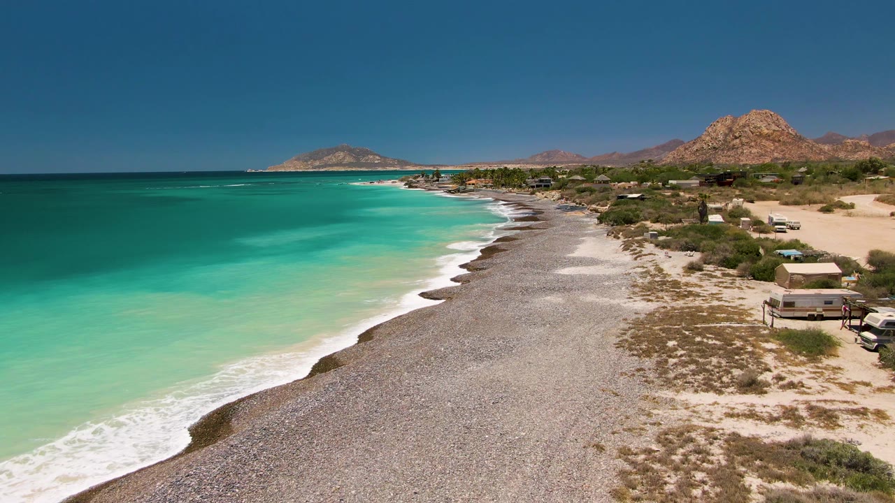 Wide drone view of turquoise sea, pebbled beach, and arid shoreline.