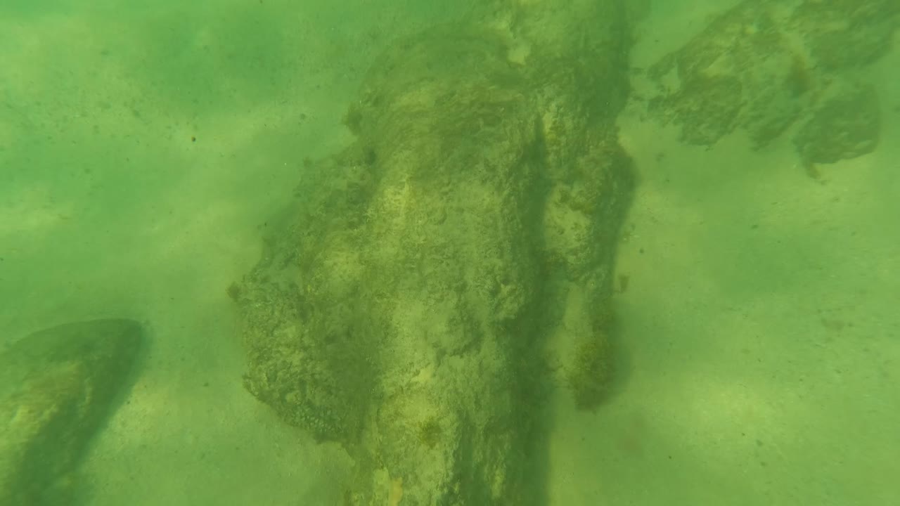 Sandy ocean floor in Los Barriles, Baja California Sur, with green water, large rock shapes near the right side and top edge, faint fish forms, and no visible people.