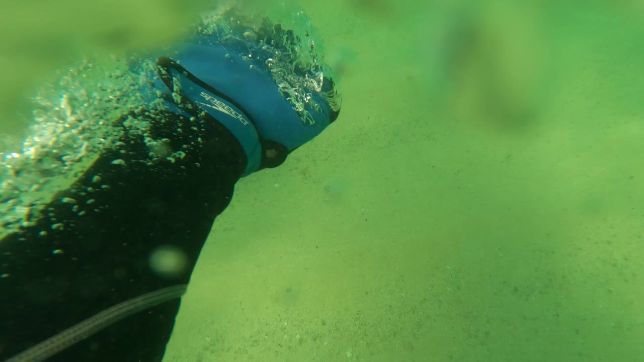 A snorkeler's arm with a blue wristband moves through green water above the sandy seabed in Los Barriles, Baja California Sur, creating bubbles near the upper right of frame.