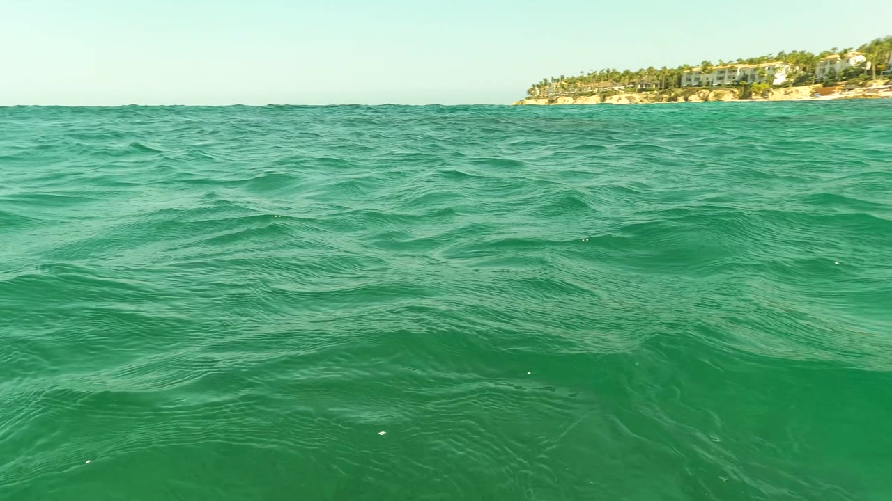 Open green-turquoise water fills most of the frame near Los Barriles, Mexico, with a low horizon and a distant developed shoreline at the far right and no visible people.