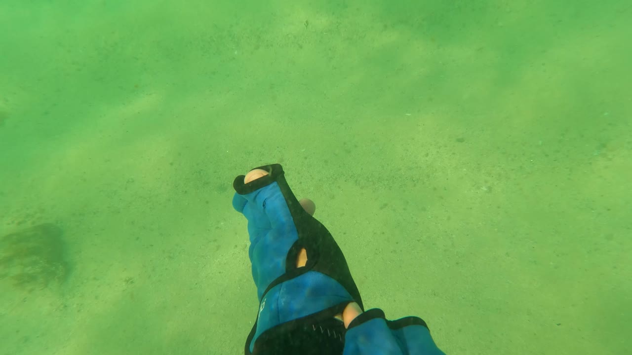 Two blue and black dive gloves are visible near the lower edge above green-lit sand in Los Barriles, Baja California Sur, with no visible face.