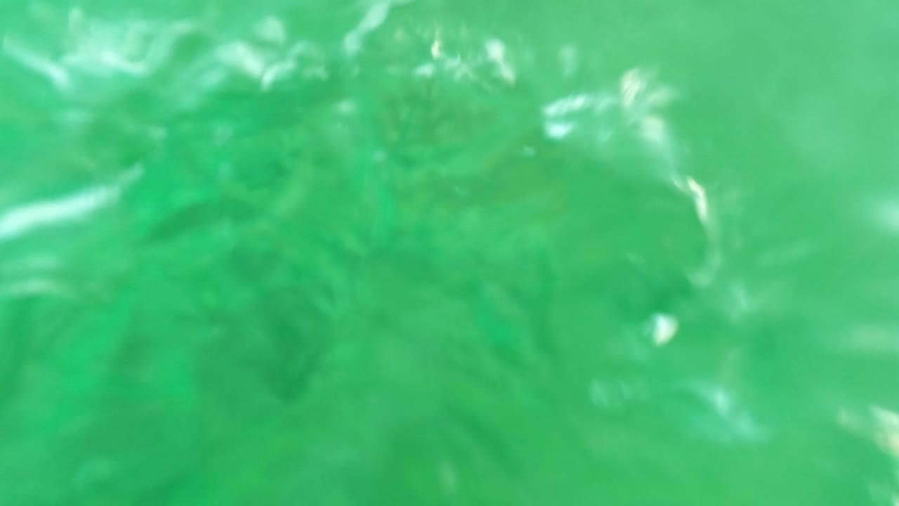 The camera looks up through green water toward a rippled surface with bright light refraction in Los Barriles, Baja California Sur, and no visible people.