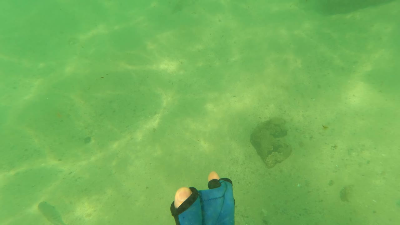A blue and black glove sits near the lower right edge of the Los Barriles underwater frame, with open green water, sandy seabed, darker rocks, and no visible people.