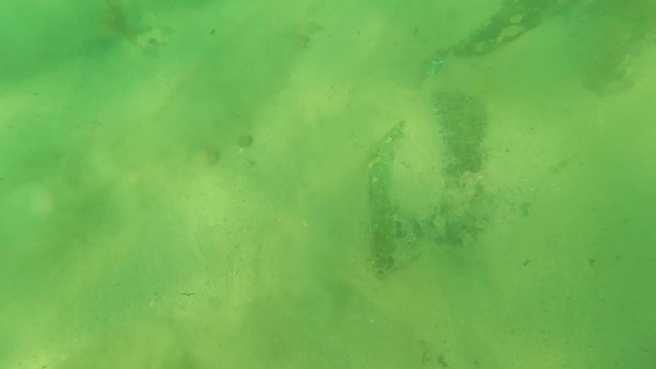 Second underwater frame in Los Barriles, Baja California Sur, Mexico, with green-tinted water, pale sand, irregular rock patches, and no visible people.