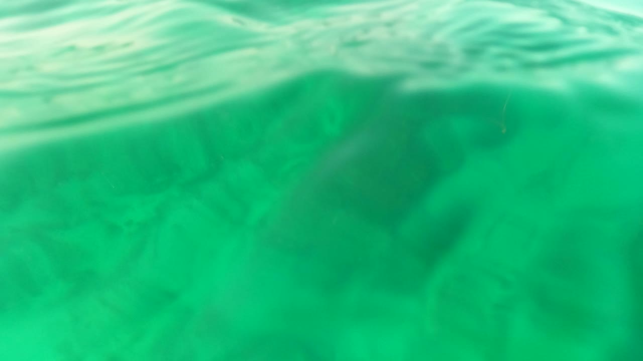 Turquoise underwater view in Los Barriles with the water surface crossing the top of frame, faint rocks beneath, and no visible people.