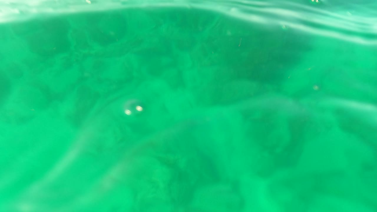 Close underwater surface ripples over green water with rocks below.