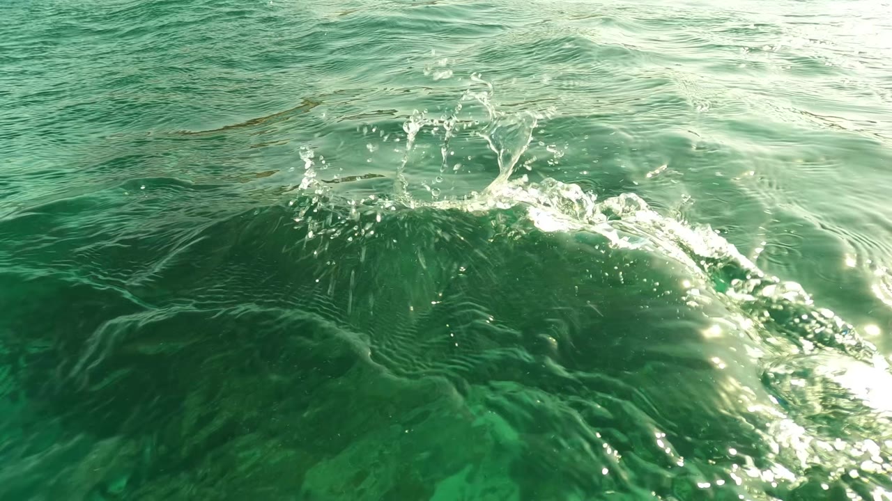 Green ocean surface in Los Barriles, Baja California Sur, with ripples, bright reflections, and a small splash near the center, no visible people.