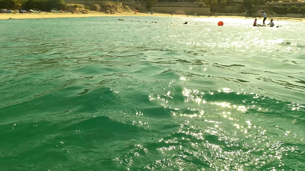 Sunlit surface water in Los Barriles, Baja California Sur, Mexico, with green ripples filling the foreground, a sandy beach strip behind it, distant swimmers, and a red buoy near shore.