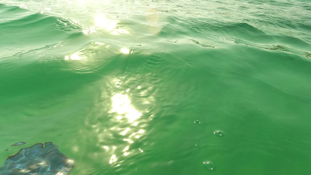 A bright wave crest and sun reflection sit over green water near the Los Barriles shoreline, with a blue gloved hand partly visible at the lower edge.