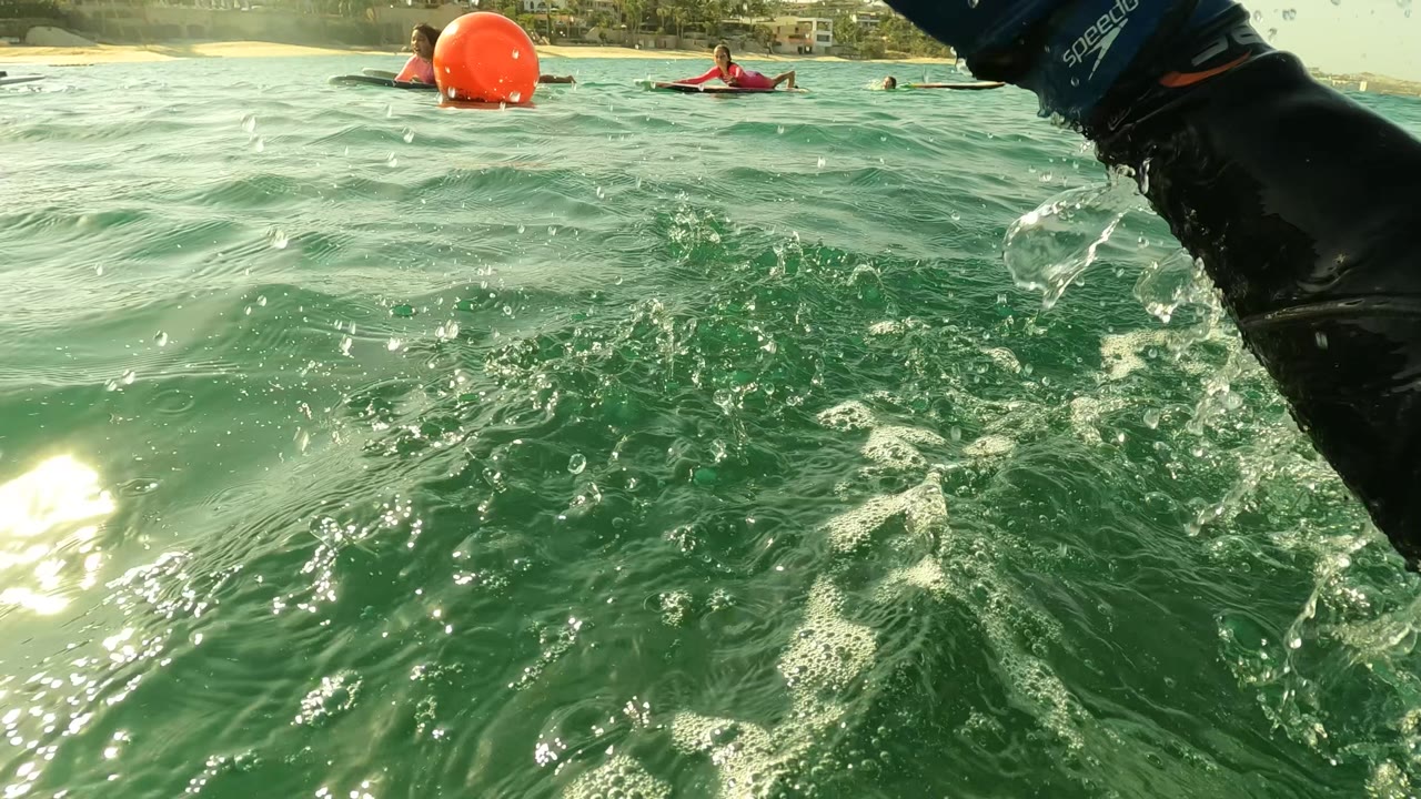 Water-level view in Los Barriles, Baja California Sur, with green ocean chop, a red buoy, distant people or watercraft near shore, and a dark wetsuit arm entering the right edge.