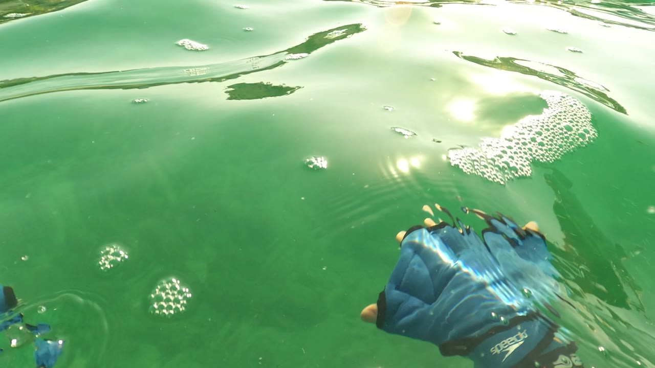 Green underwater surface view from Los Barriles, Baja California Sur, with bright glare, small bubbles, and a blue-gloved hand entering at the right edge.