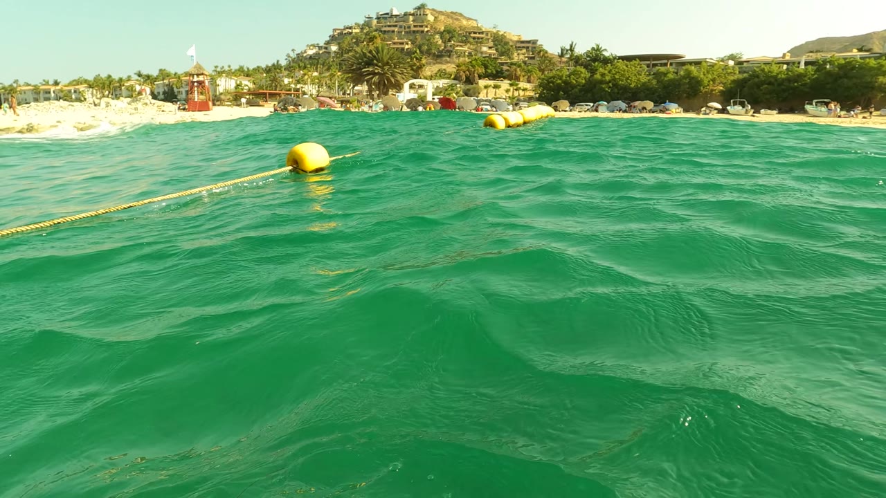 Water-level view in Los Barriles, Baja California Sur, with turquoise waves in front, yellow buoys on a rope, sandy beach, palms, buildings, and no visible people in this frame.