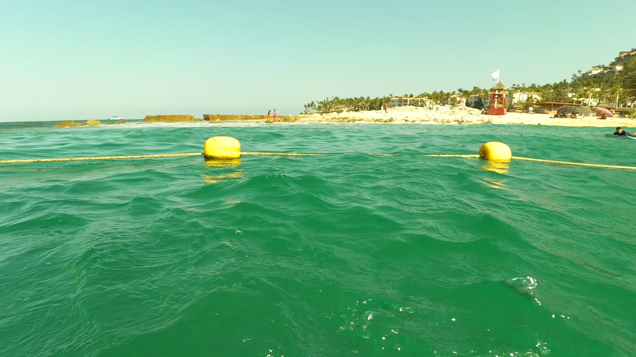 The Los Barriles shoreline recedes to the right as a yellow buoy and rope cross the green-blue water, with a distant swimmer near the edge of frame and beach structures beyond.