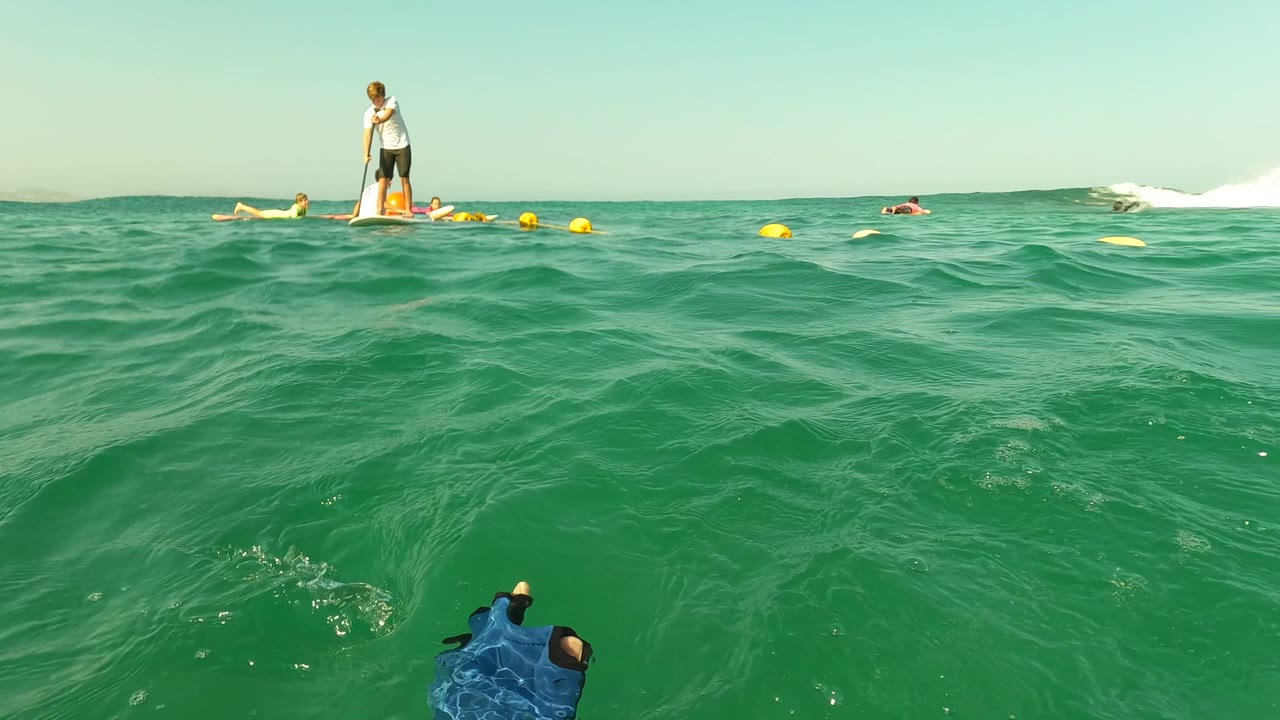 Water-level frame in Los Barriles, Baja California Sur, showing green ocean water, a buoy line, swimmers, and paddleboarders spread across the horizon.