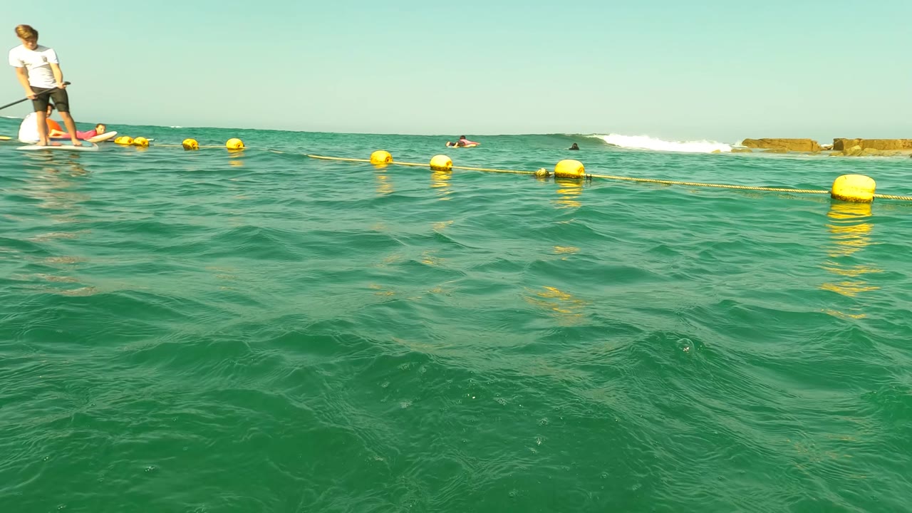 Paddleboarder standing near yellow buoys with swimmers in the surrounding ocean.