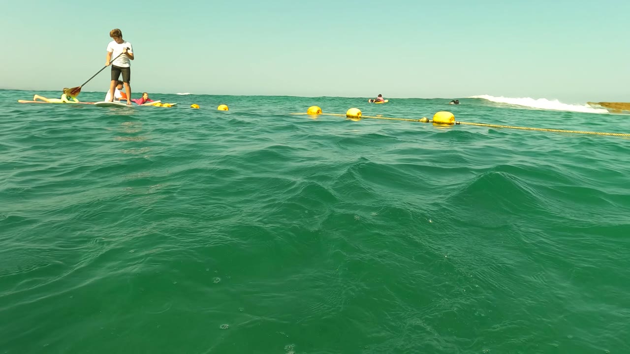 Los Barriles ocean surface fills most of the frame, with a paddleboarder at the upper right, swimmers near the horizon, and yellow buoys stretching across the water.
