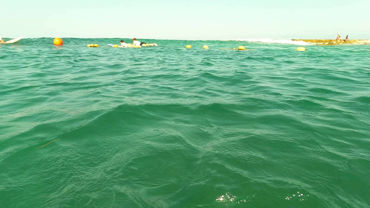 Water-level view in Los Barriles, Baja California Sur, Mexico, with green ocean waves, yellow buoys, distant swimmers, and a pale clear sky.