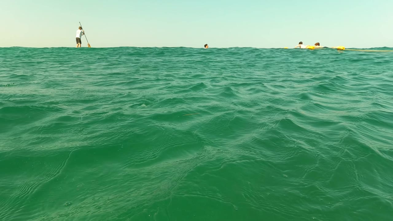 Green ocean water rises across the foreground in Los Barriles, with a paddleboarder standing near the horizon and small swimmers and yellow buoys behind the wave line.