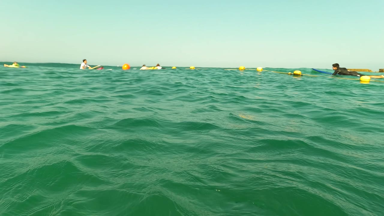 Water-level view in Los Barriles, Baja California Sur, with green waves in the foreground and distant swimmers floating near yellow buoys under clear sky.