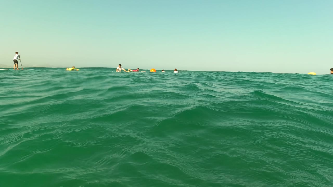 Swimmers and floaters spread across green ocean water beside yellow buoys.