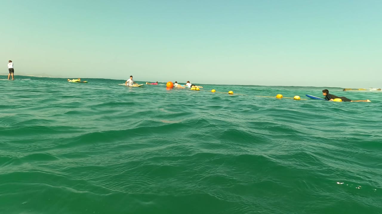 Water-level view in Los Barriles with textured green waves filling most of the frame and small distant figures floating near yellow buoys along the horizon.