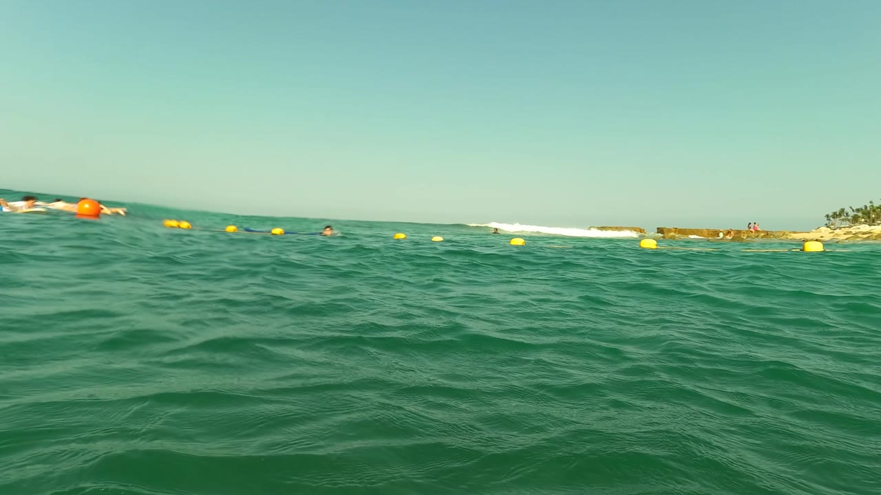 Water-level view in Los Barriles, Baja California Sur, Mexico, with swimmers spread across the horizon line, yellow buoys floating in green water, and bright sky above.
