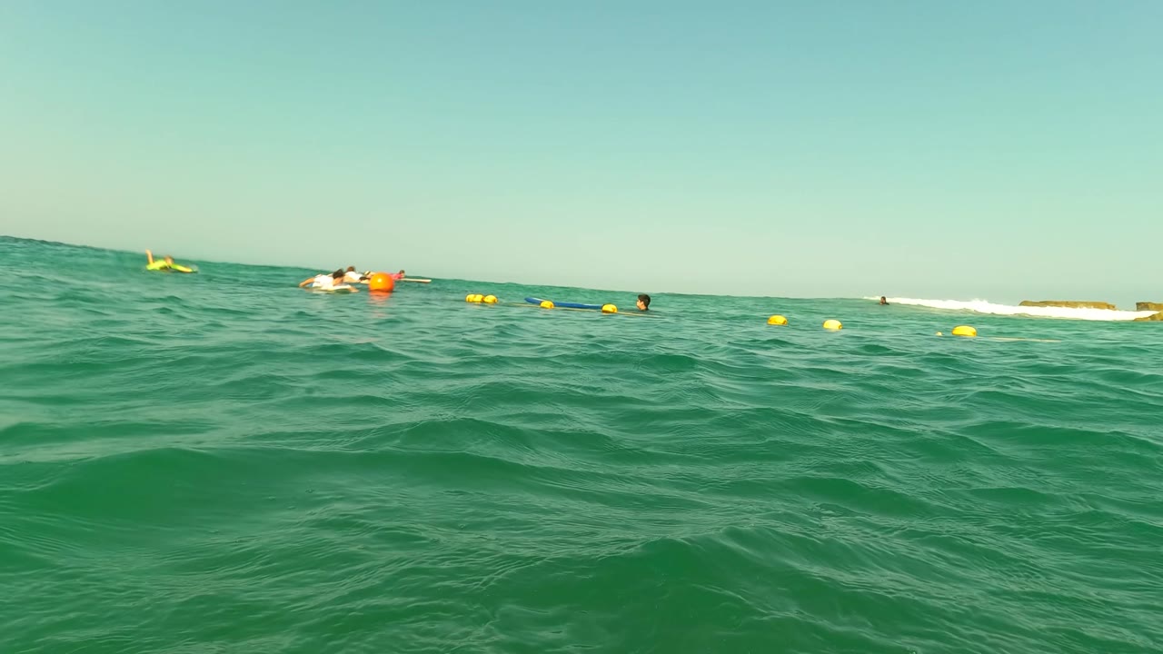 Green waves rise in the foreground while yellow buoys and distant swimmers sit near a low shoreline in Los Barriles, Baja California Sur, Mexico.