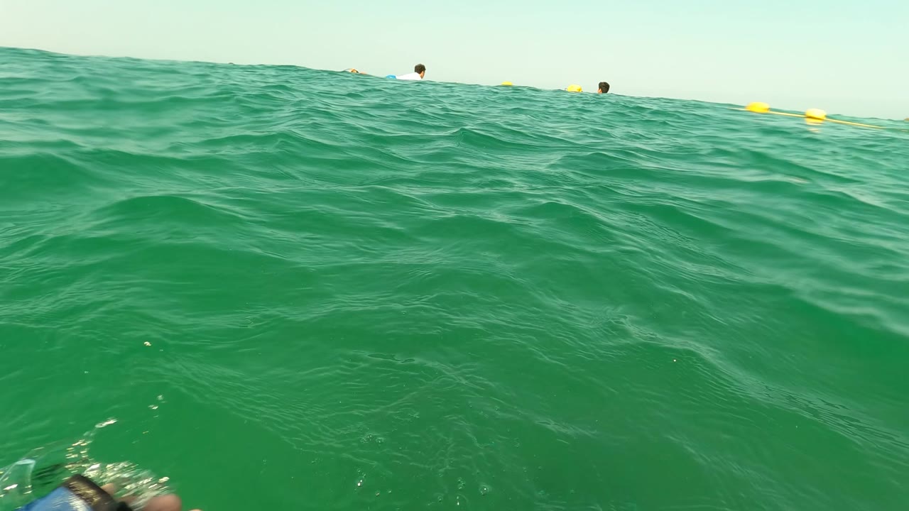 Water-level view in Los Barriles, Baja California Sur with green waves in the foreground, small swimmers near a yellow buoy line, and a faint shoreline on the horizon.