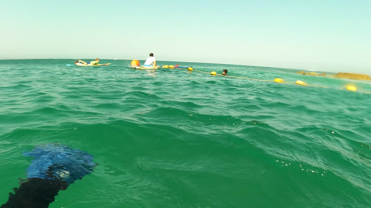A low waterline frame in Los Barriles, Baja California Sur shows yellow and orange buoys, several distant swimmers, and a small craft or floating object near the horizon.