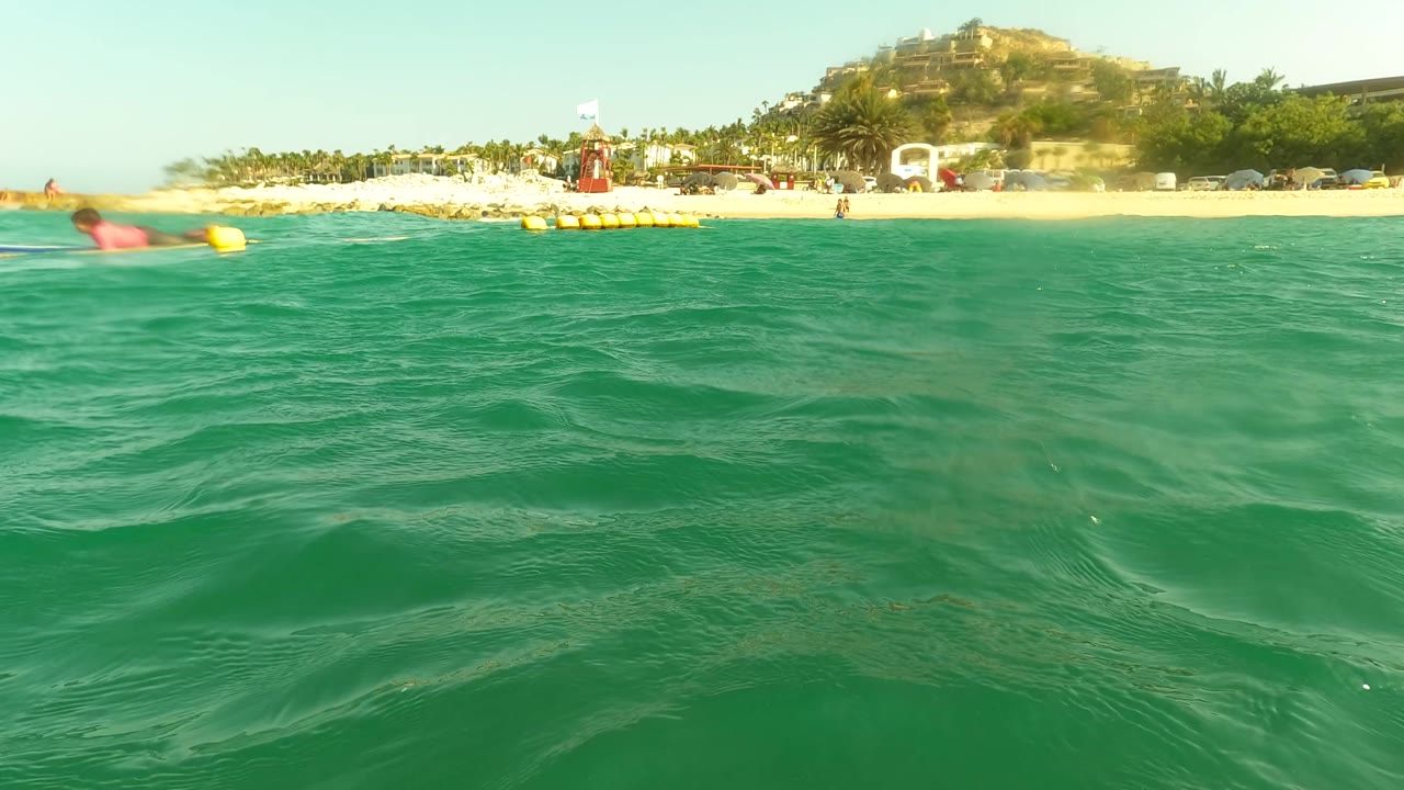 Water-level view in Los Barriles, Baja California Sur, Mexico, with turquoise waves in the foreground, yellow buoys near shore, beachgoers on the sand, and hillside buildings in the background.
