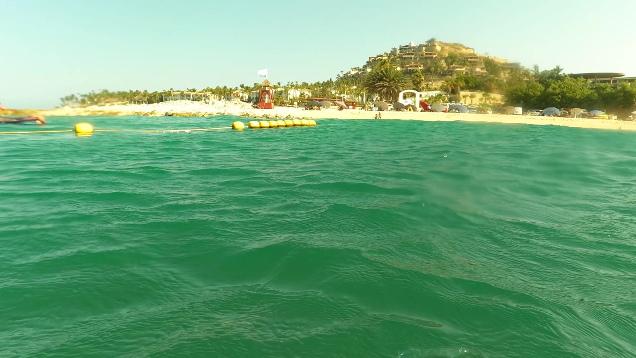 Ocean surface with a beach, yellow buoys, and distant shoreline buildings.
