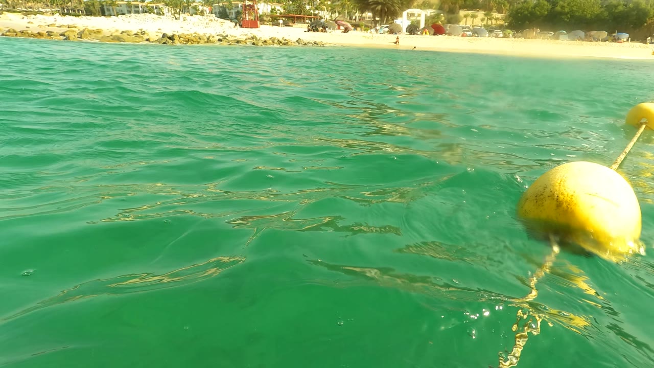 The water-level view in Los Barriles includes a yellow buoy at the right edge, turquoise surface ripples, and sandy beach with distant shoreline detail.