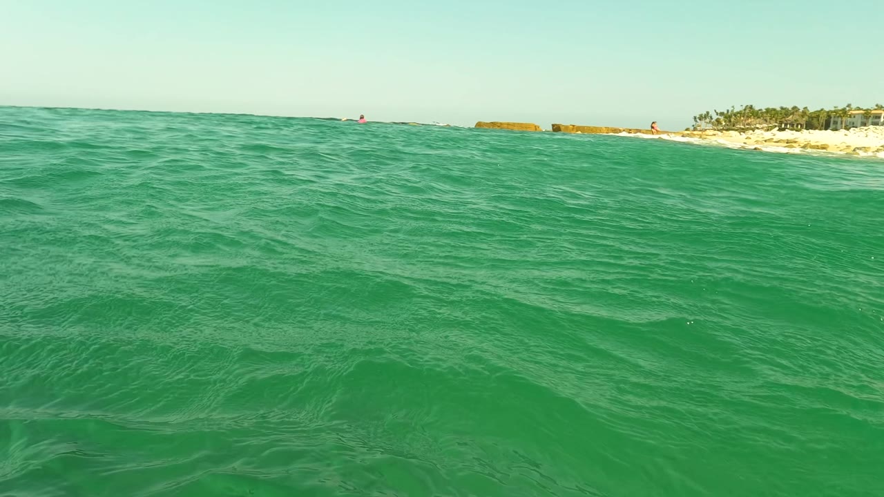Water-level frame in Los Barriles showing turquoise waves, a sandy shoreline with palms, and tiny distant beachgoers along the right side.