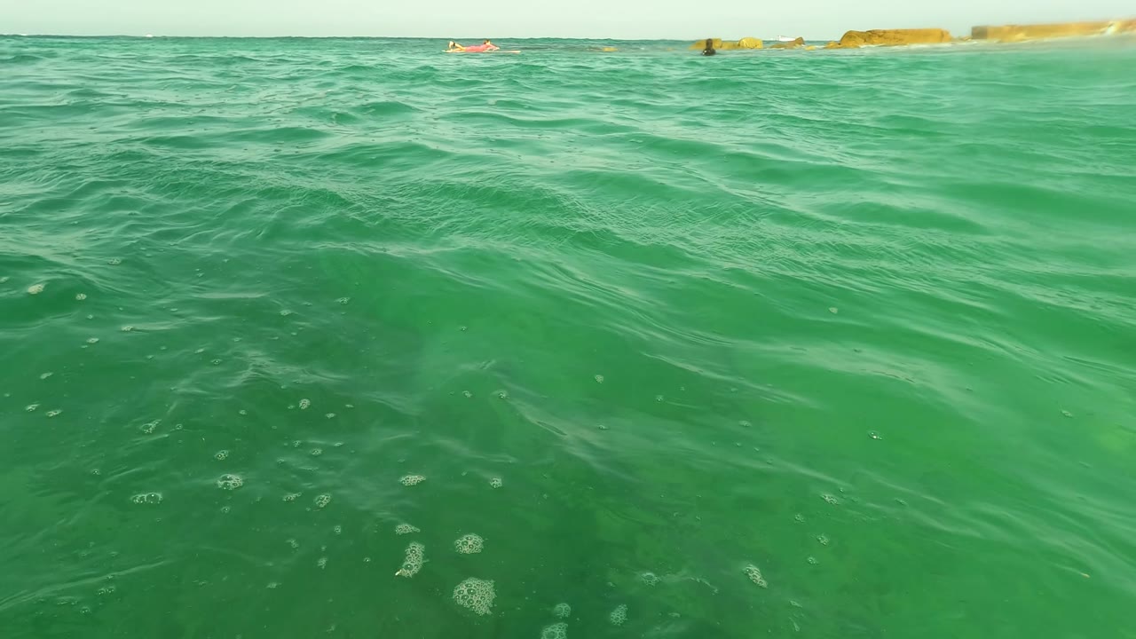 Clear green ocean surface at Los Barriles, Baja California Sur, with ripples in the foreground, a low horizon, distant rocks, and small distant swimmers visible.