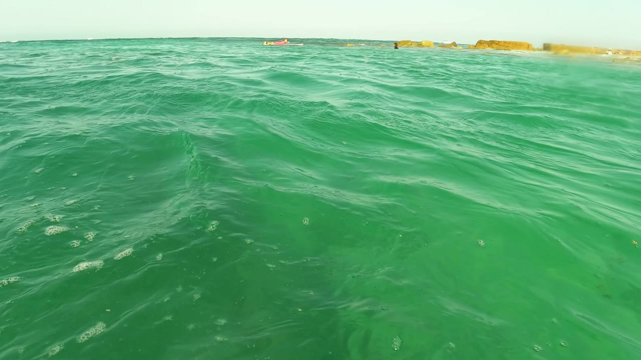 A water-level frame from Los Barriles, Baja California Sur, shows clear green waves, a small distant swimmer near the horizon, and low rocks in the background.