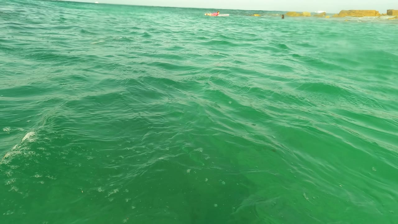 Rippling green sea fills the Los Barriles frame, with low background rocks and tiny distant swimmers along the horizon line.