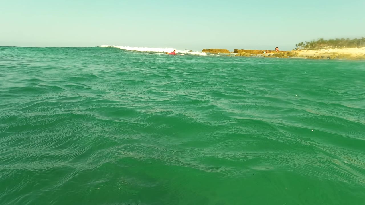 Water-level view in Los Barriles, Baja California Sur, with turquoise waves in the foreground, a rocky sandy shoreline in the distance, and small distant people near the beach.