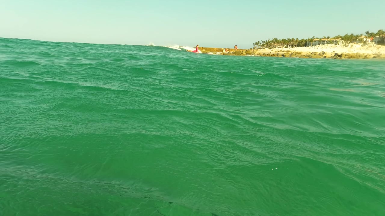 In Los Barriles, the frame shows a broad turquoise wave surface, low beach rocks, palm trees along the coast, and small distant people on the sandy shoreline.