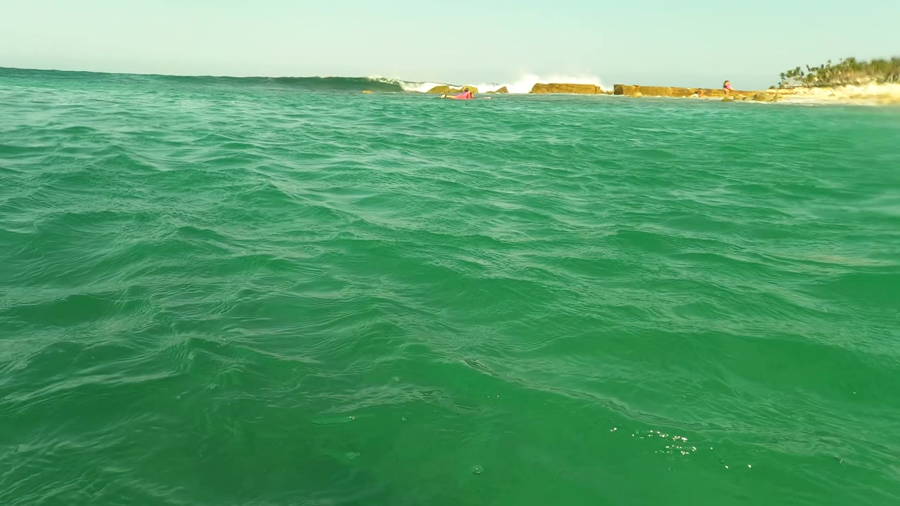 Water-level view of turquoise ocean at Los Barriles, Baja California Sur, with rippled foreground water, white surf, a sandy shoreline, and tiny distant people near the beach.