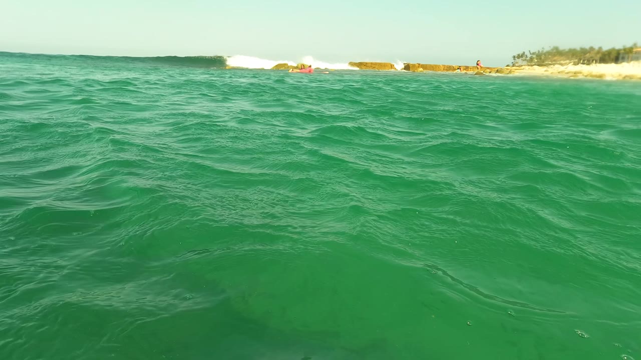Green ocean ripples with white surf breaking near a distant beach.