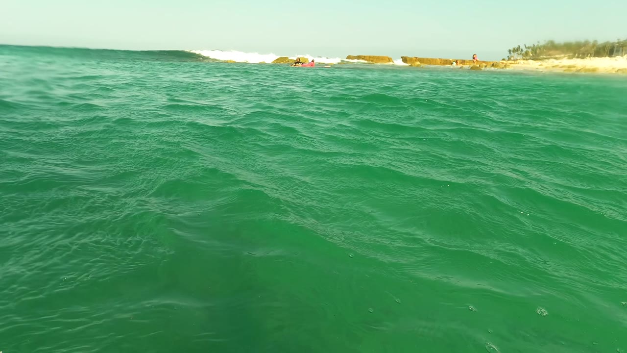 A low waterline view at Los Barriles, Baja California Sur shows choppy turquoise surface texture, whitewater near shore, a sandy beach, and very small distant people.