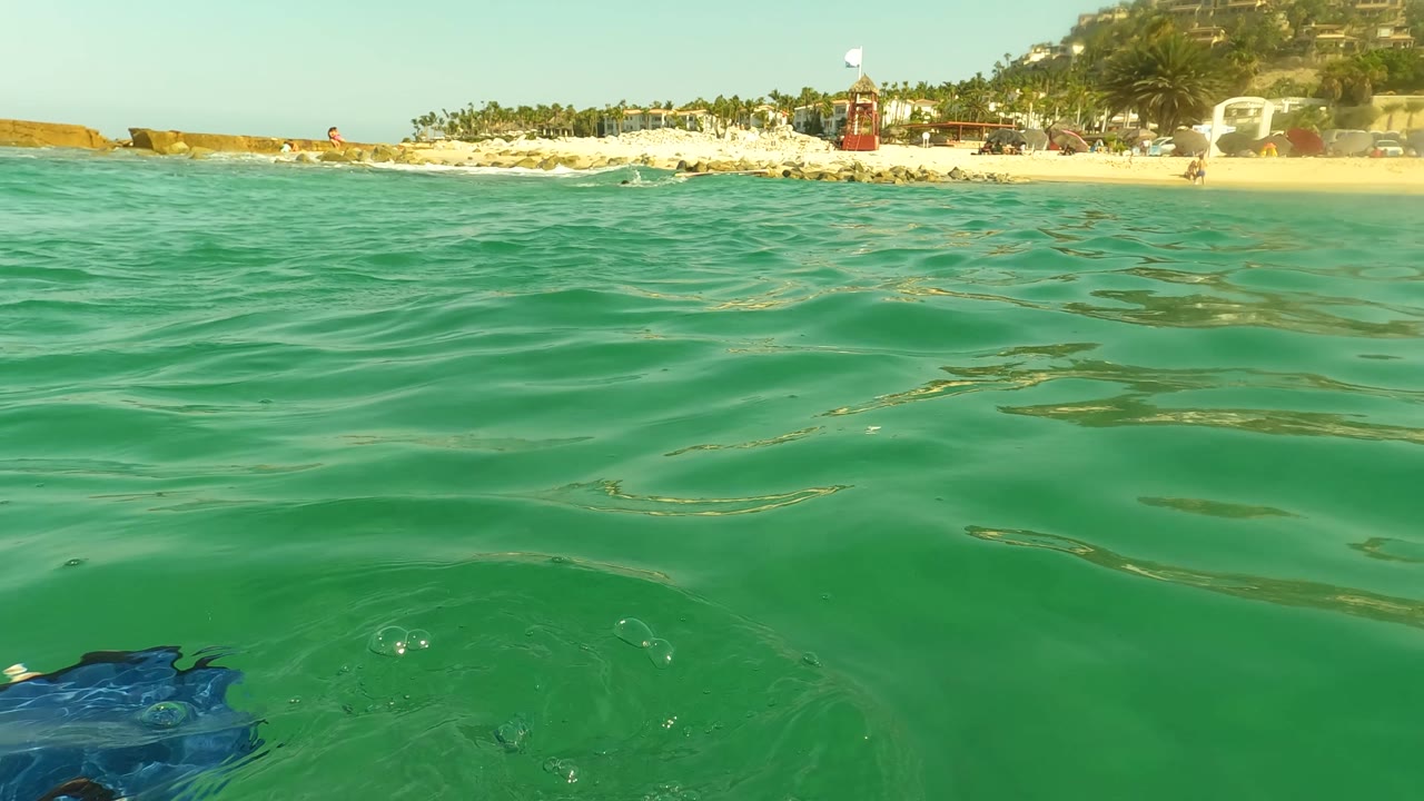 Water-level view in Los Barriles, Baja California Sur, with green rippled ocean in front, sandy beach and palms on the horizon, and no visible people.