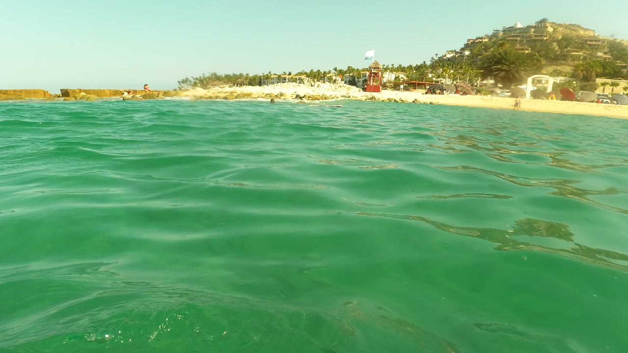 A wider water-level moment shows the green ocean surface dominating the frame, with the Los Barriles beach and palm-lined shore set high on the horizon and no visible people.