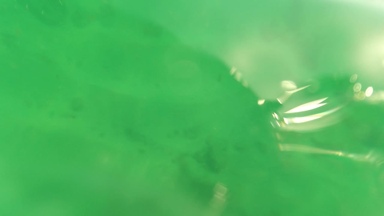 Green underwater frame from Los Barriles with a sandy bottom, bright surface reflection near the upper right, a small fish-like shape, and no visible people.