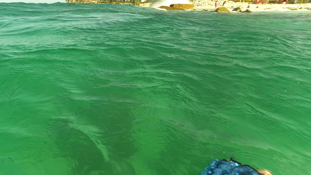 Surface-level green ocean water near Los Barriles, Baja California Sur, with a distant sandy beach along the top edge and a partial swimmer shape at the bottom, no visible people identifiable.