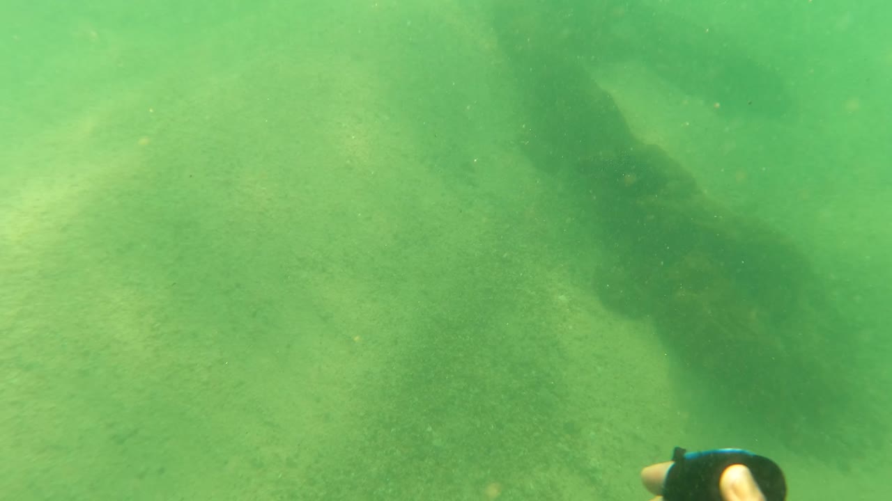 Underwater view in Los Barriles, Baja California Sur with a darker algae-covered form stretching through the center over pale sand and no visible people.