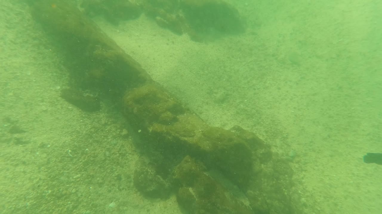 A long algae-covered submerged structure crosses the sandy seabed in green water near Los Barriles, Baja California Sur, with no visible people.