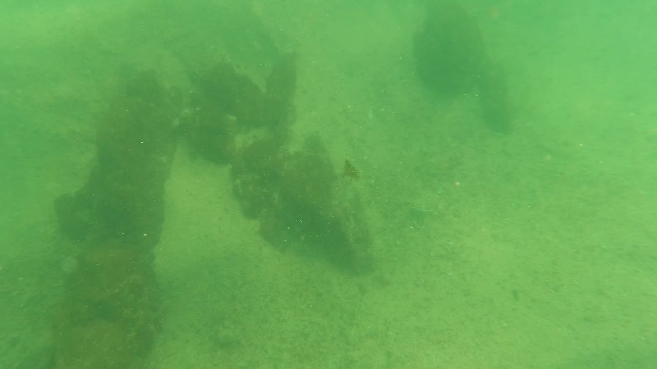 A darker section of the rusted submerged structure fills the center of the Los Barriles underwater frame, with pale sand visible behind it and no visible people.